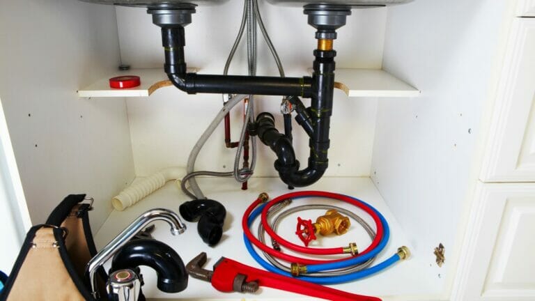 Assortment of professional plumbing tools and fittings arranged in front of sink pipes within a kitchen cupboard, symbolizing the work of a qualified plumber.