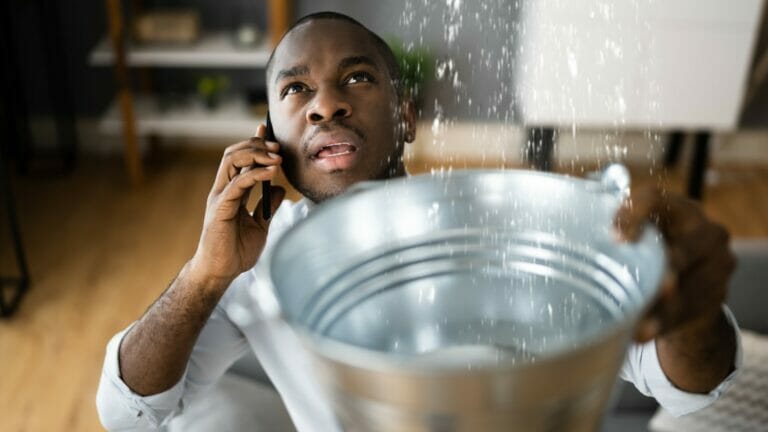 A concerned man holding a bucket to collect water leaking from the ceiling while calling a plumber on his cellphone, highlighting the urgency of finding a reliable commercial plumber to address such issues promptly and efficiently.