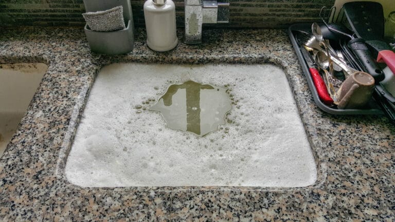 Overflowing soapy water on a granite kitchen countertop near utensils, indicating a clogged sink.