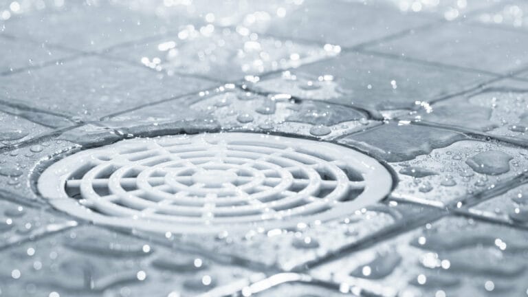 A close-up of water droplets on a tiled floor surrounding a circular shower drain, illustrating the importance of keeping shower drains unclogged for smooth water flow.