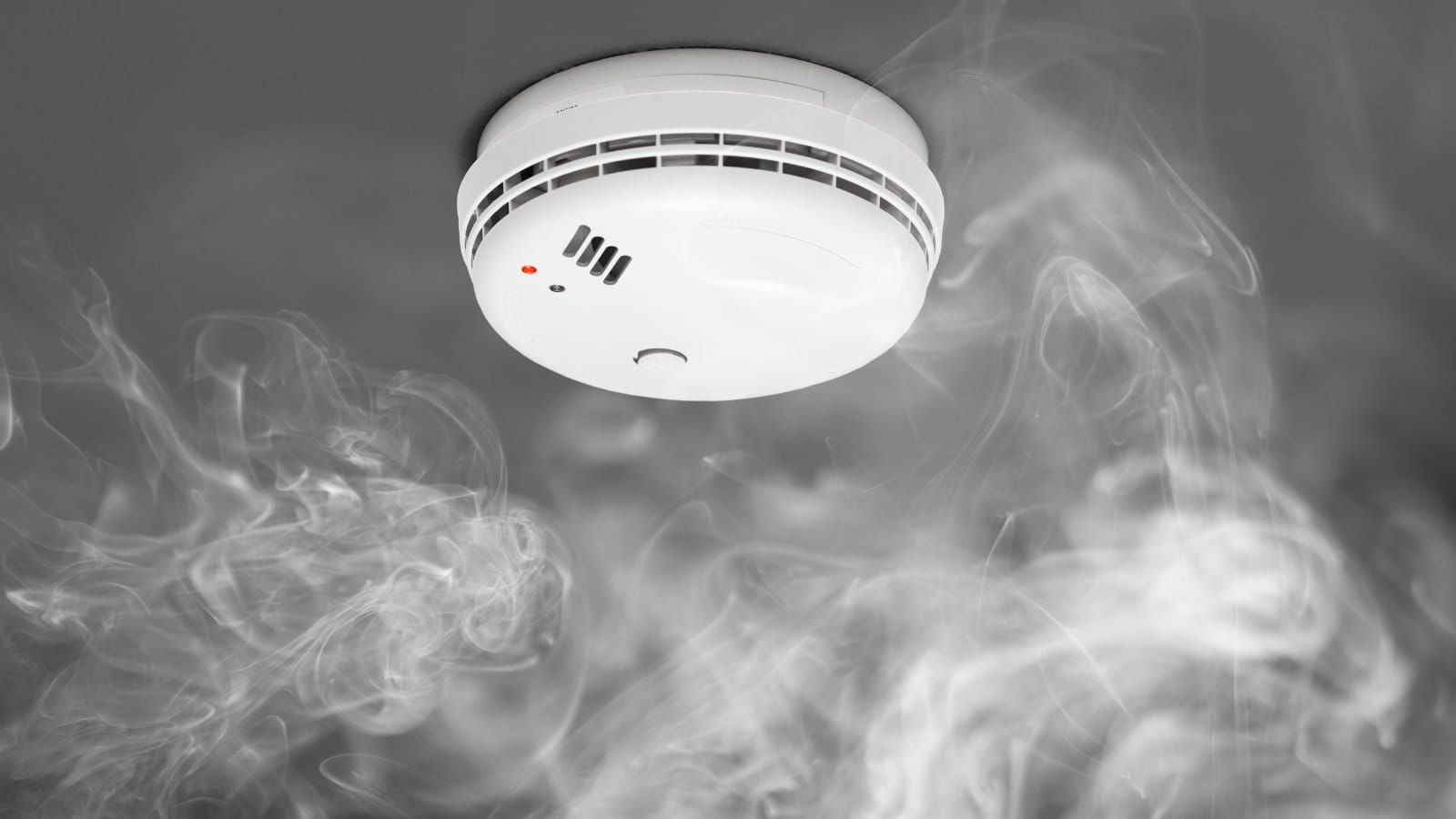 Smoke alarm activated with visible smoke against a gray background, illustrating the importance of fire safety and the functionality of smoke detectors in a home setting.