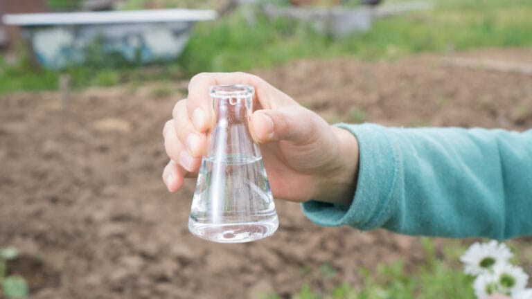 A hand holding a clear vial of water with a blurred natural background, representing water quality testing for safe drinking water.