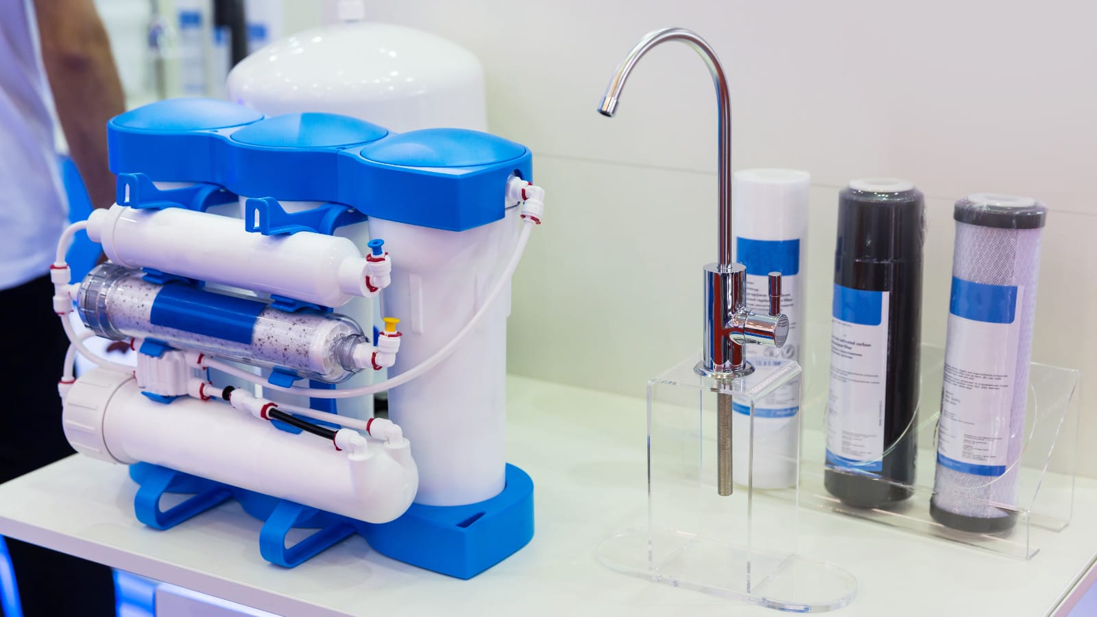 A modern home water filtration system on display with various filter stages, including sediment and charcoal filters, connected by pipes to a chrome faucet, demonstrating residential water purification technology.