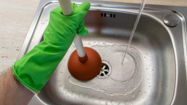 A person's hand clad in a bright green rubber glove is using a red plunger in a stainless steel kitchen sink, with water flowing from the faucet, depicting a common plumbing issue of a clogged drain being fixed.