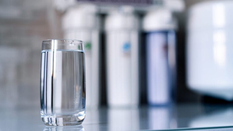 A glass of clear water in sharp focus on a kitchen counter with a blurred background featuring water filtration systems, symbolizing the purity and cleanliness of water obtained through home water filtration, as discussed in the benefits of such systems