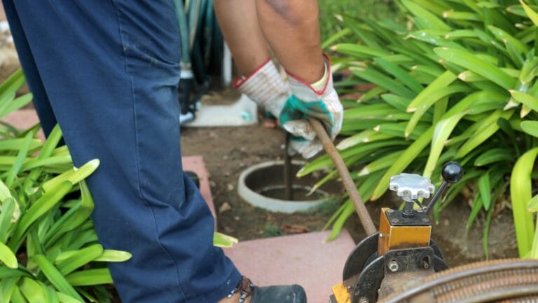 A sewer-cleaning plumber uses a sewer snake to clear a blockage in a sewer line, surrounded by green plants, indicating outdoor maintenance work.