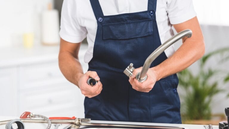 Midsection of a man in a navy blue apron holding a disassembled kitchen faucet, preparing to install or repair it, representing a DIY plumbing scenario.