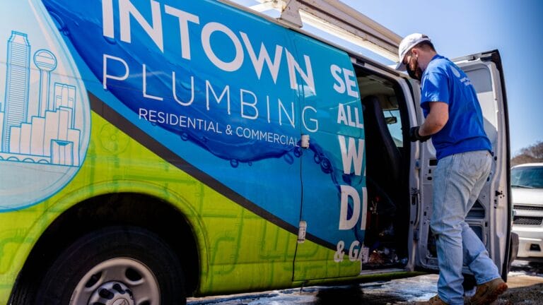 Plumber in blue uniform reaching for tools in a brightly wrapped Intown Plumbing Services van, indicating professional plumbing assistance for residential and commercial needs.