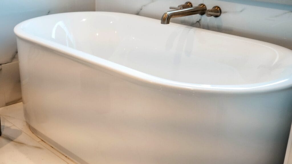 A modern white freestanding bathtub with a sleek bronze faucet, set against a marble-tiled bathroom wall.
