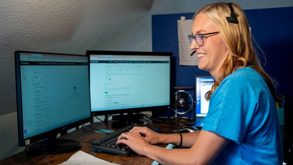 A customer service representative wearing a headset smiles while working on a computer with dual monitors, handling plumbing service requests.